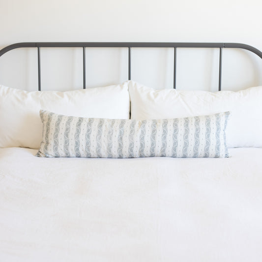 Long rectangular lumbar pillow with gray and white vertical striped pattern on bed with white pillows and metal headboard.