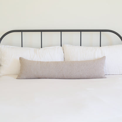 Tan and white striped lumbar pillow displayed on a bed with white pillows and metal headboard.