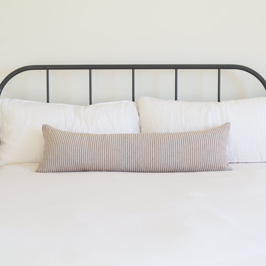 Tan and white striped lumbar pillow displayed on a bed with white pillows and metal headboard.