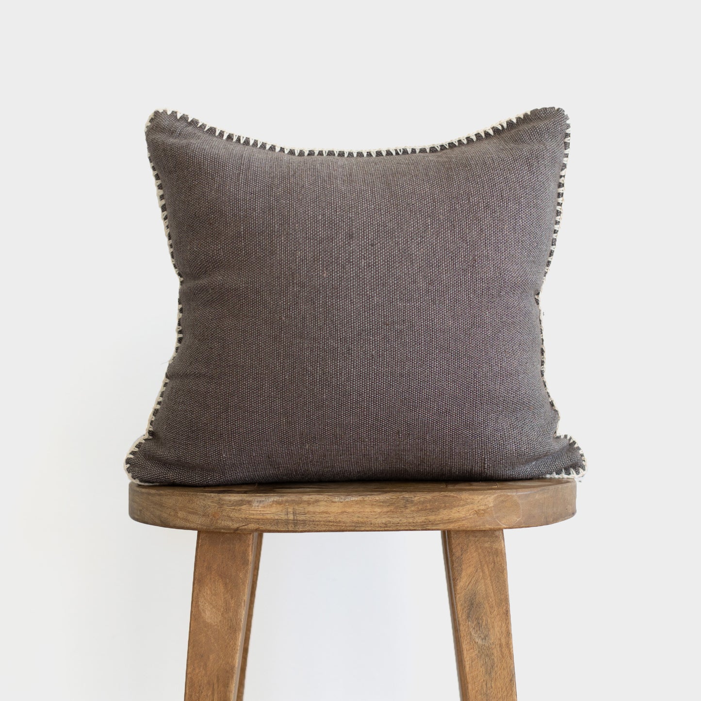 Warm charcoal cushion with scalloped edges displayed on wooden stool.