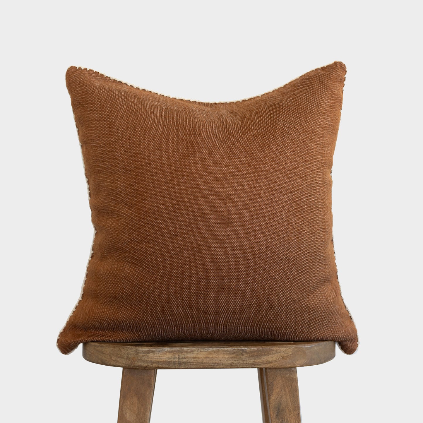 Brown throw pillow with whipstitched edges displayed on wooden stool.