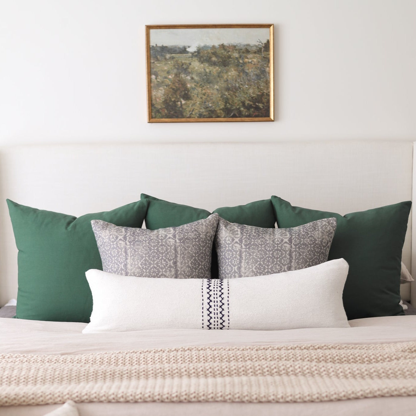 Framed landscape painting with pine wood border hung above bed with green and patterned pillows.