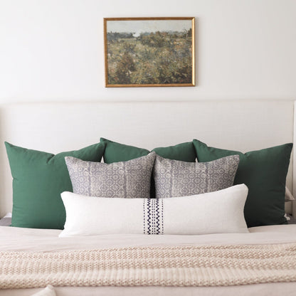 Framed landscape painting with pine wood border hung above bed with green and patterned pillows.