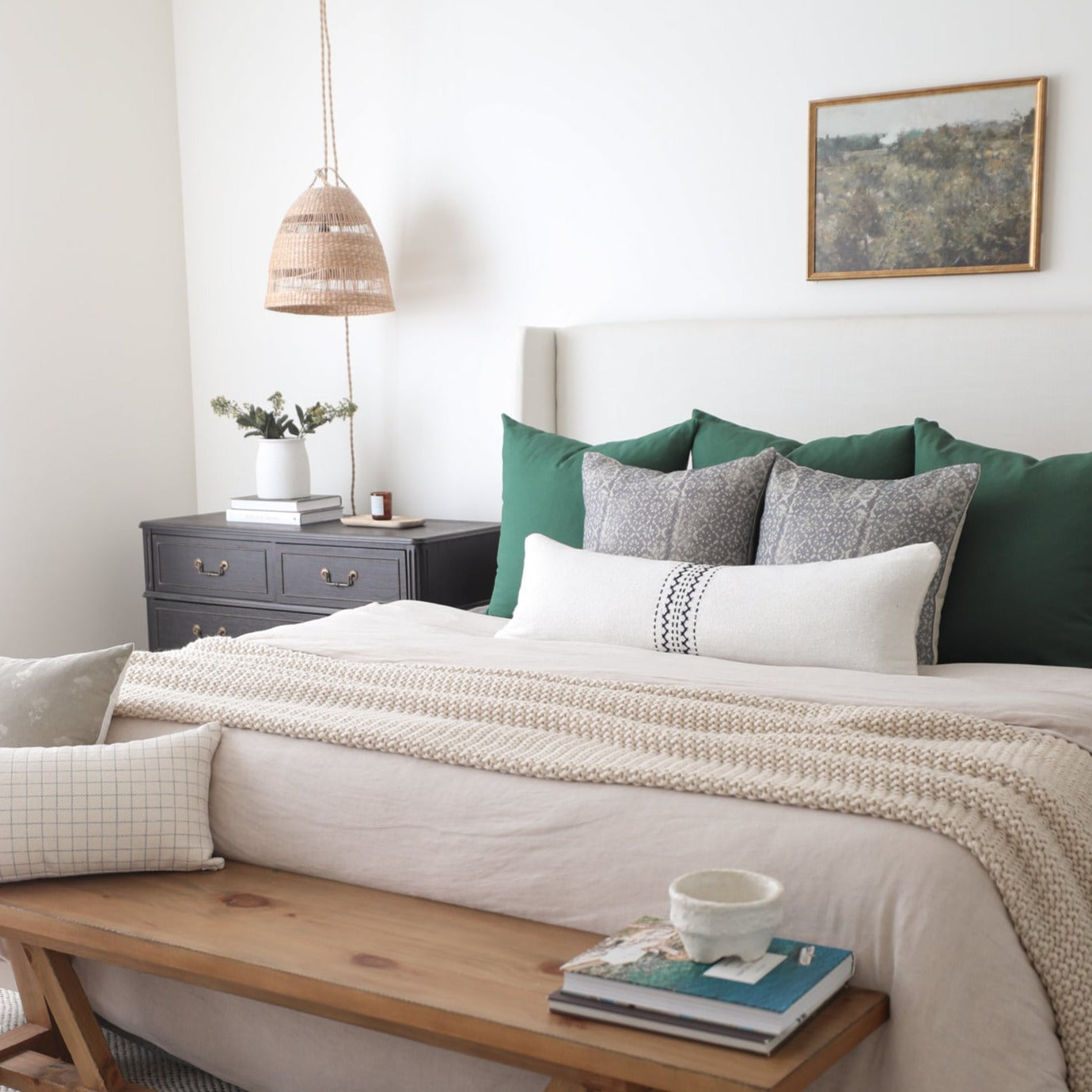 Styled bedroom with bed featuring green and gray pillows, cream bedding, wooden bench at foot, nightstand with woven pendant 