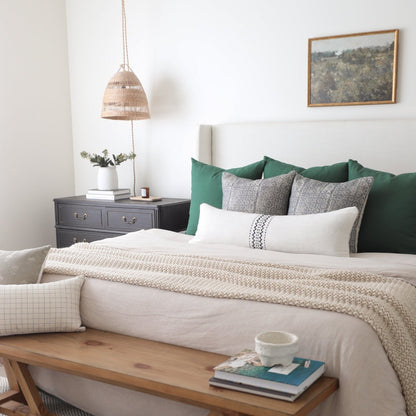 Styled bedroom with bed featuring green and gray pillows, cream bedding, wooden bench at foot, nightstand with woven pendant 