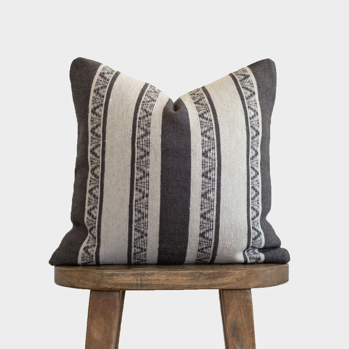 Striped throw pillow in charcoal and cream with geometric patterned bands, displayed on a wooden stool.