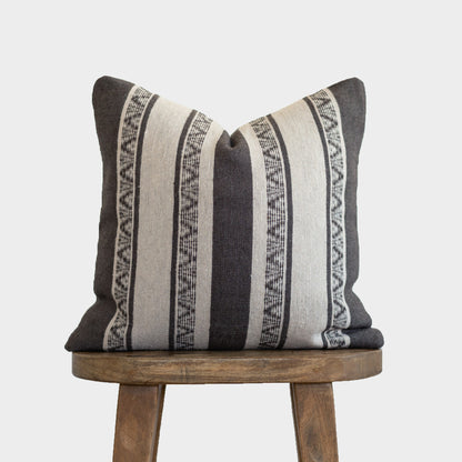 Striped throw pillow in charcoal and cream with geometric patterned bands, displayed on a wooden stool.