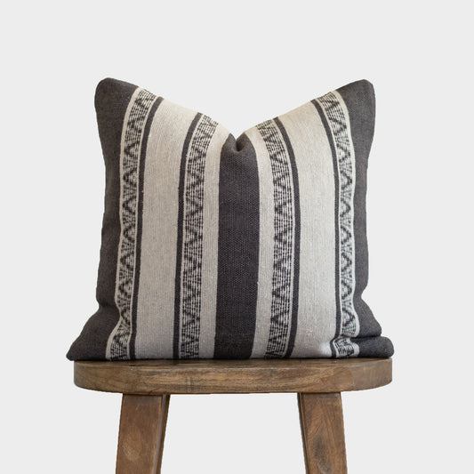Striped throw pillow in charcoal and cream with geometric patterned bands, displayed on a wooden stool.