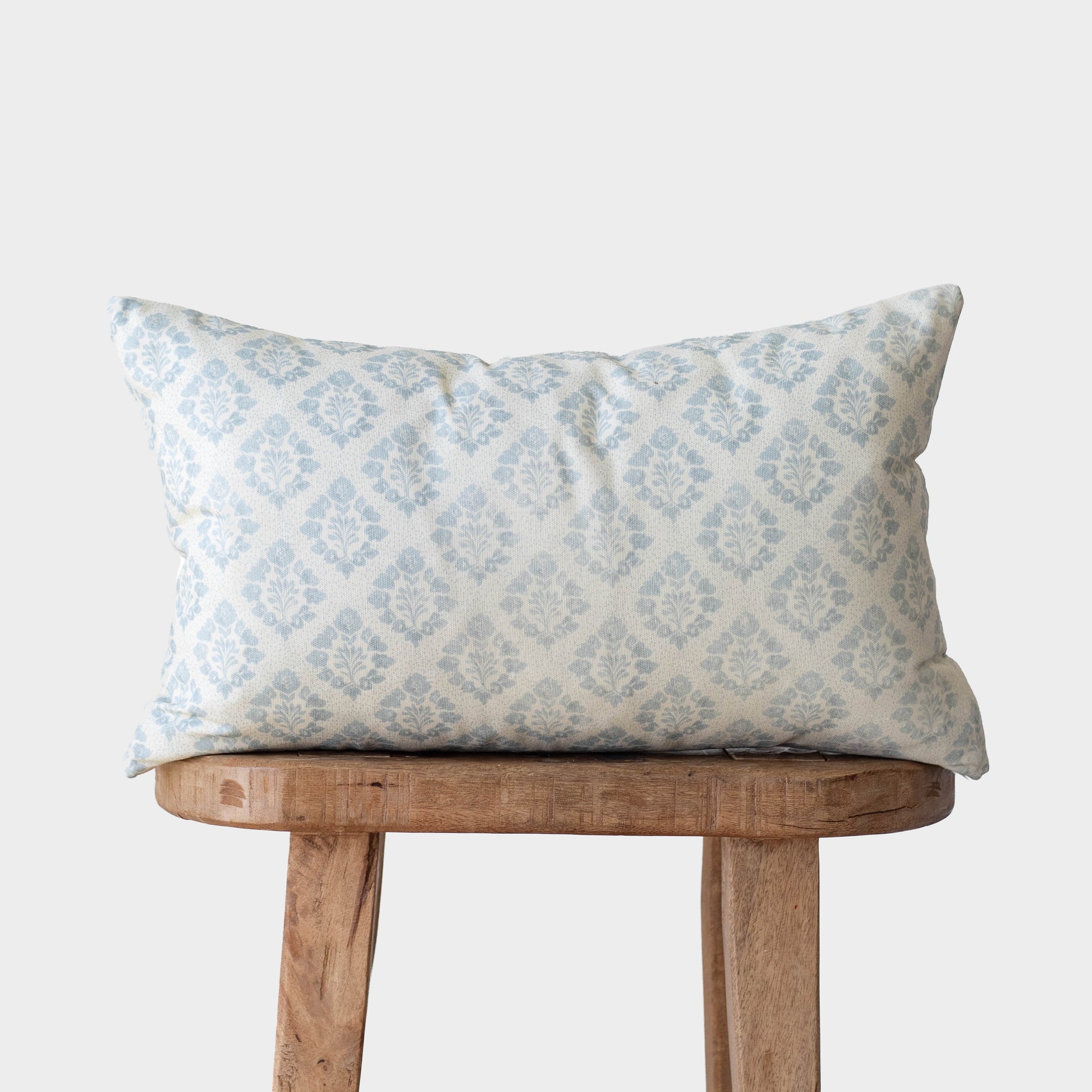 Rectangular pillow with light blue damask pattern on cream fabric, displayed on wooden stool.