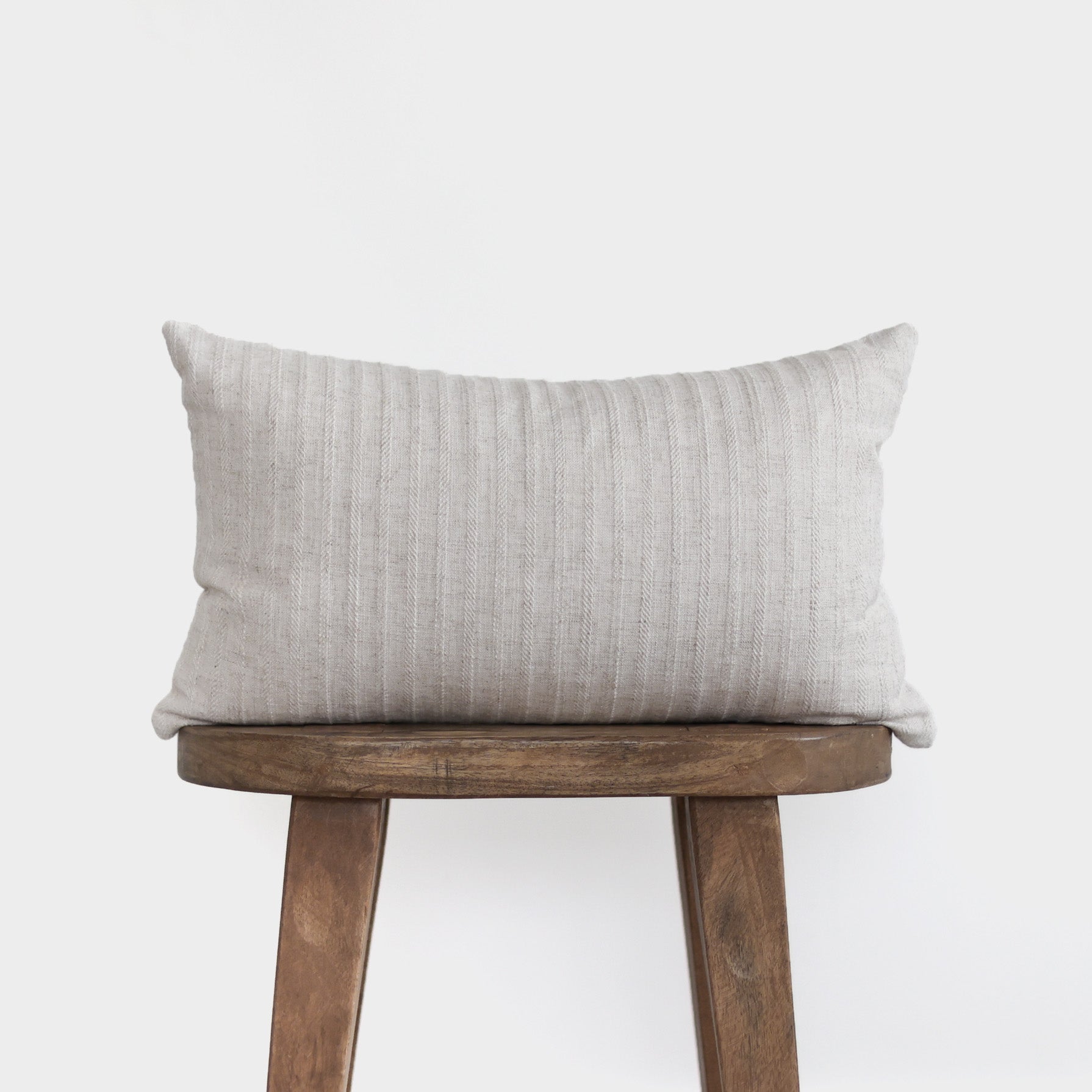 Light gray striped lumbar pillow displayed on wooden stool against white background.