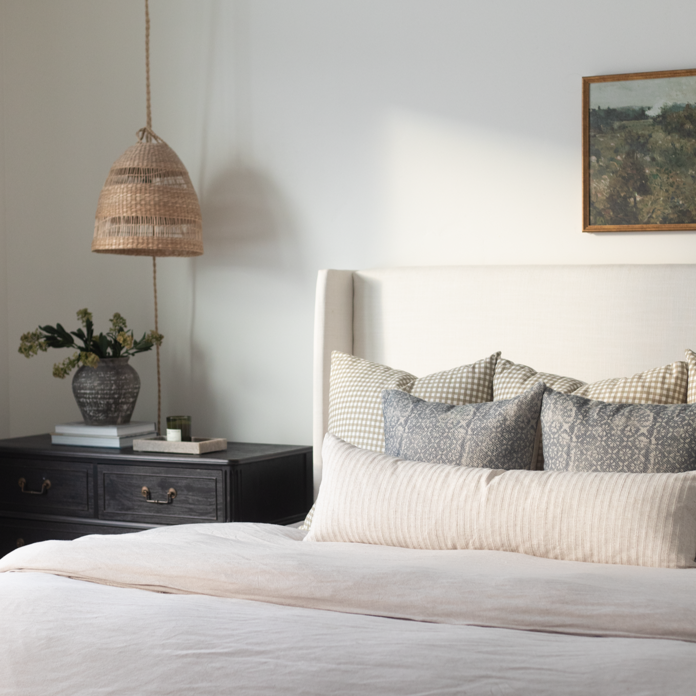 Bedroom scene with white bed, patterned pillows, woven pendant lamp, black nightstand, and framed artwork on white wall.