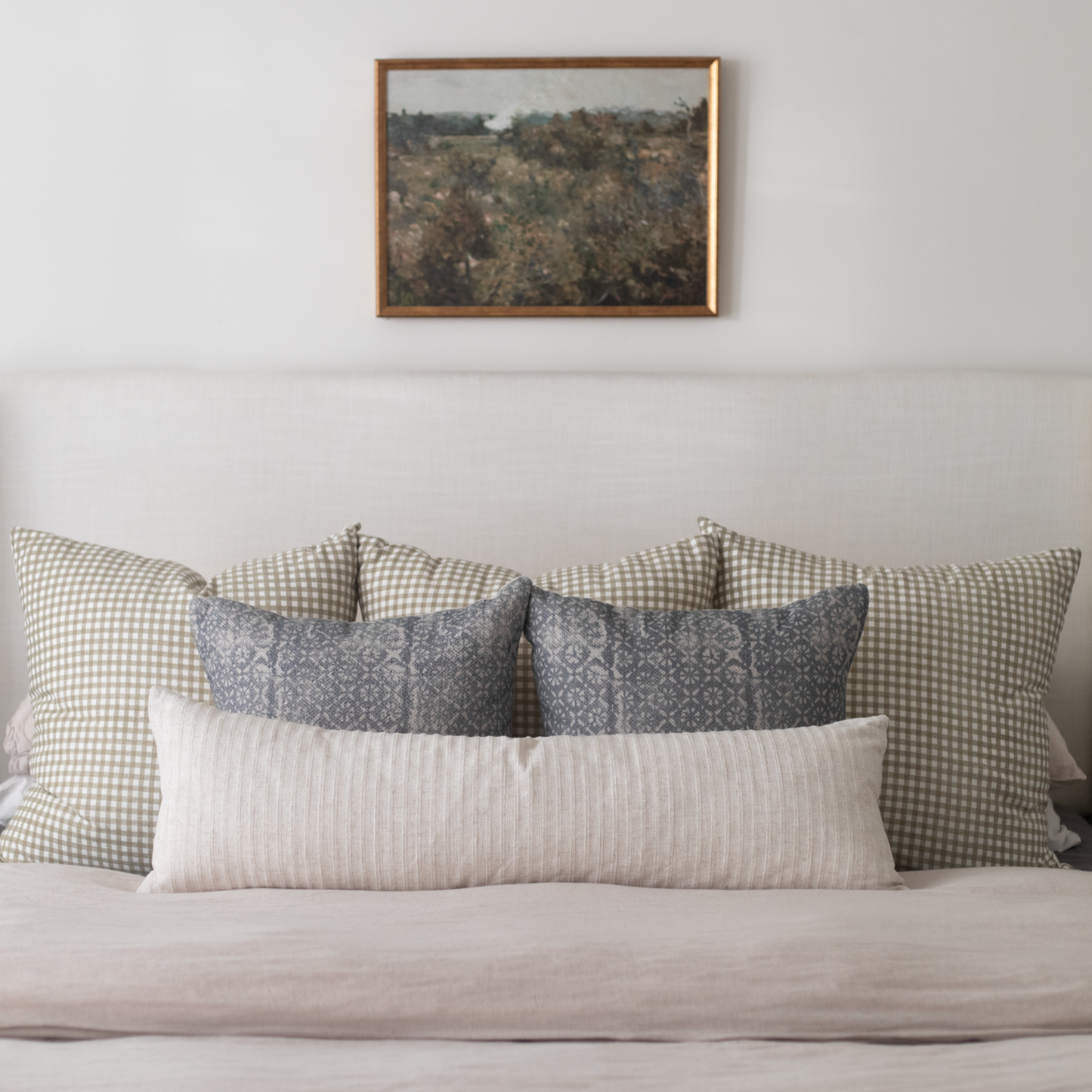 Framed landscape artwork with brown wooden frame hanging above a bed with patterned pillows.