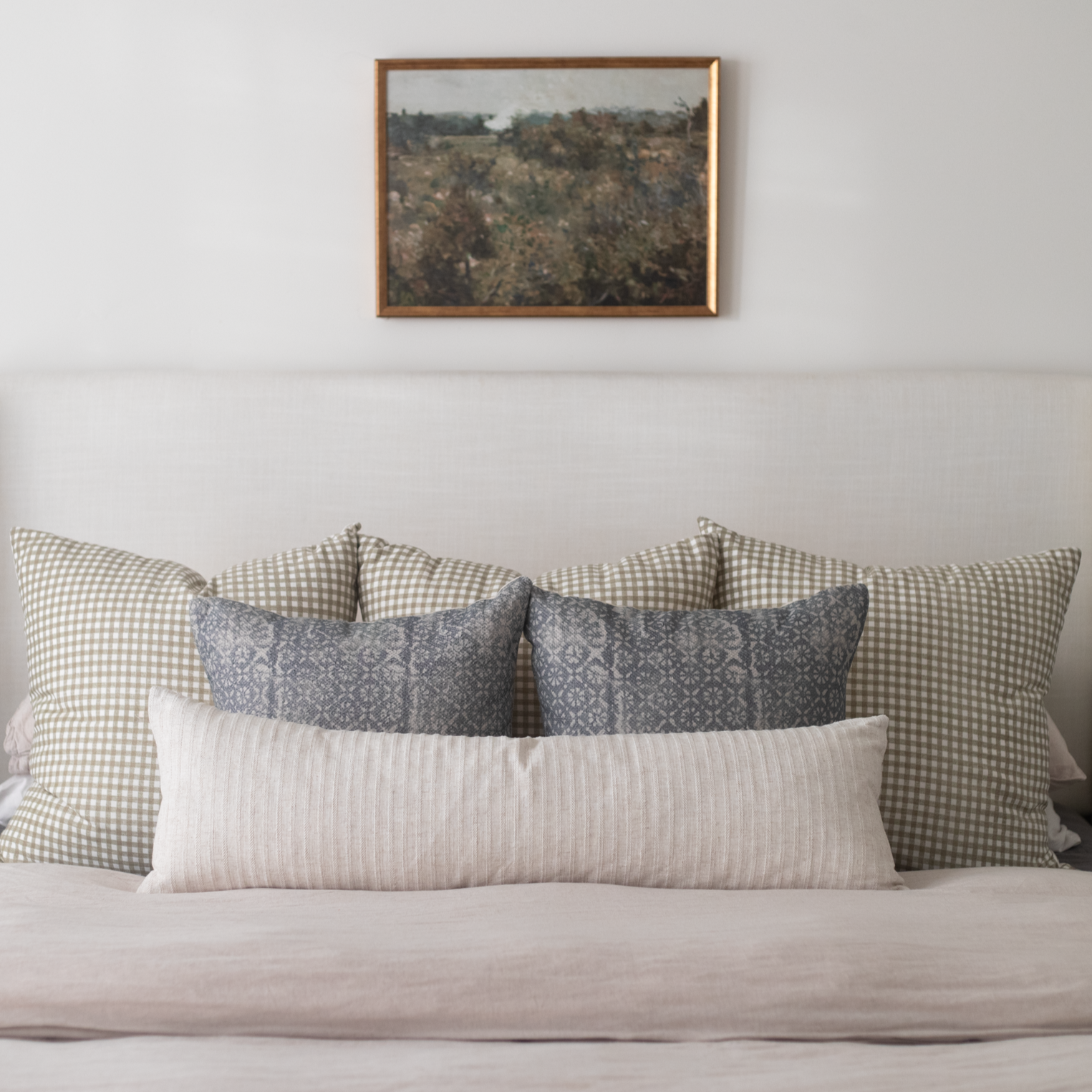 Framed landscape artwork with brown wooden frame hanging above a bed with patterned pillows.