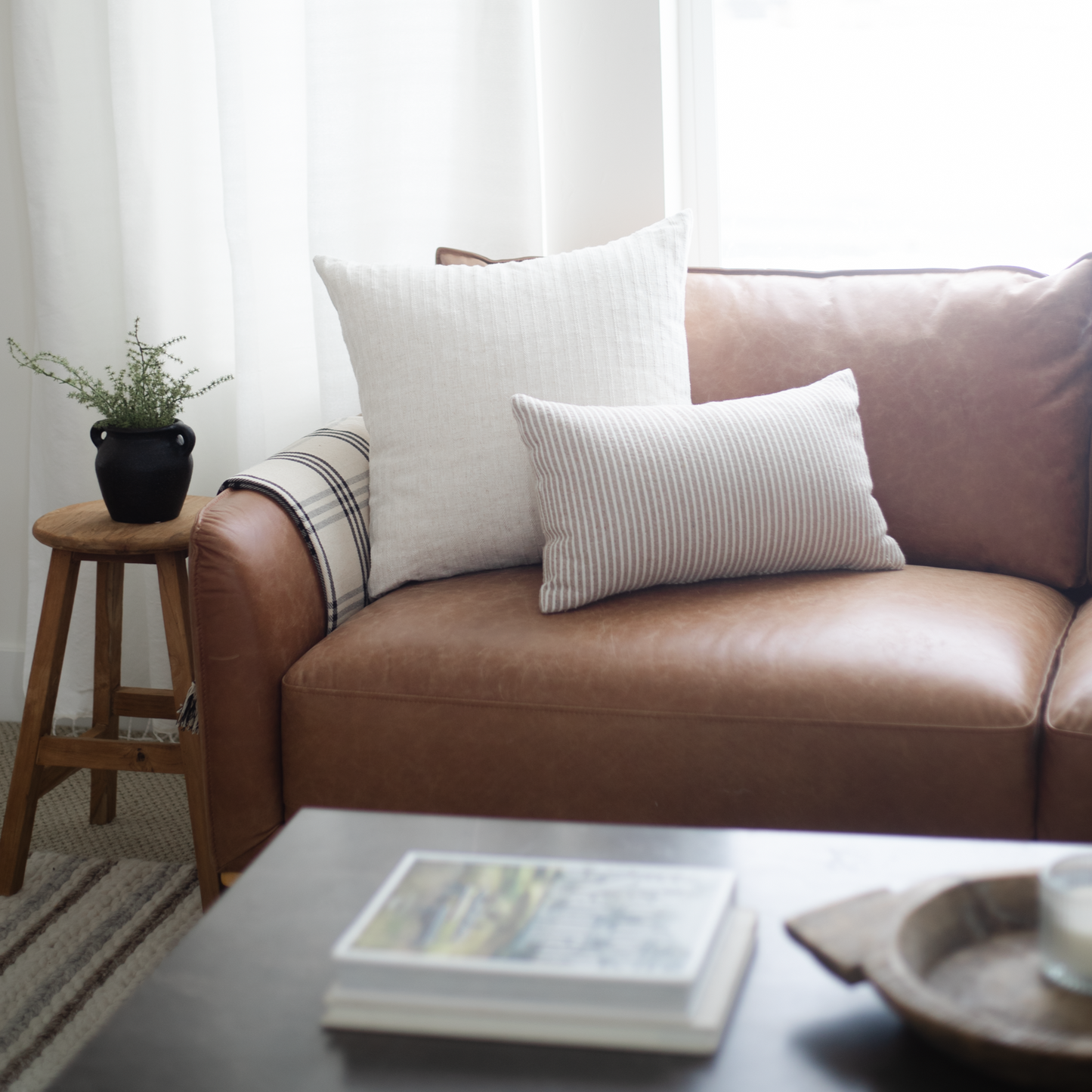 Brown leather sofa with white and mauve ribbed pillows, wooden side table with black planter, and striped throw blanket.