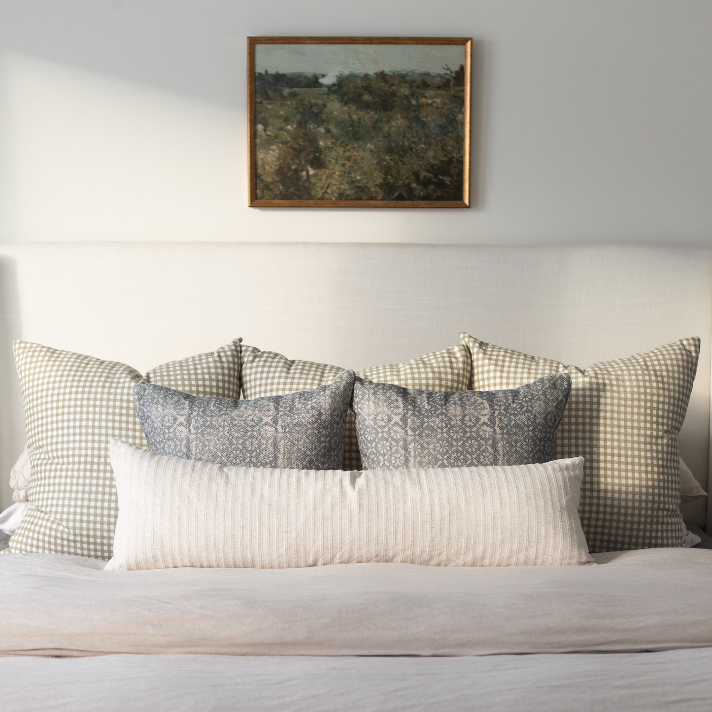 Framed landscape painting with brown wood frame hanging above bed with patterned pillows.