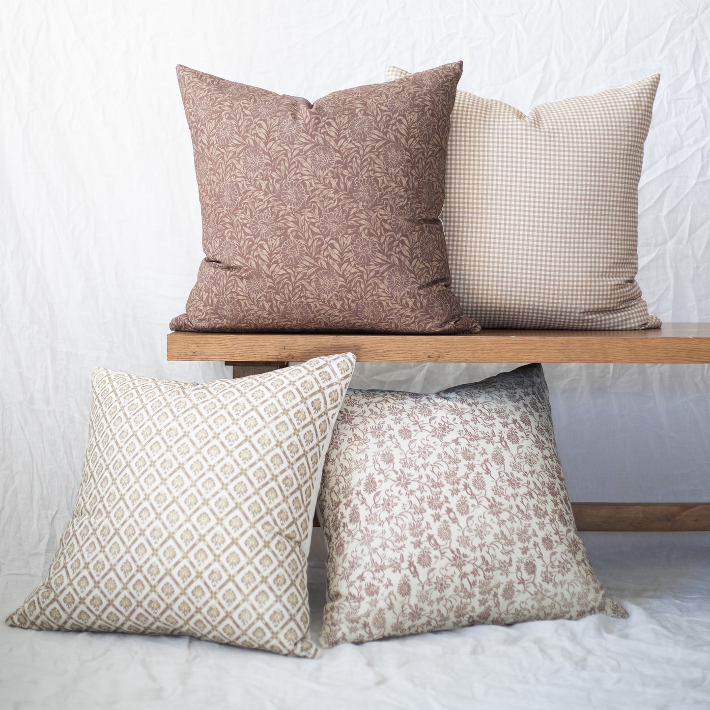 Four decorative pillows in taupe and cream with floral, gingham, and geometric patterns displayed on wooden bench.