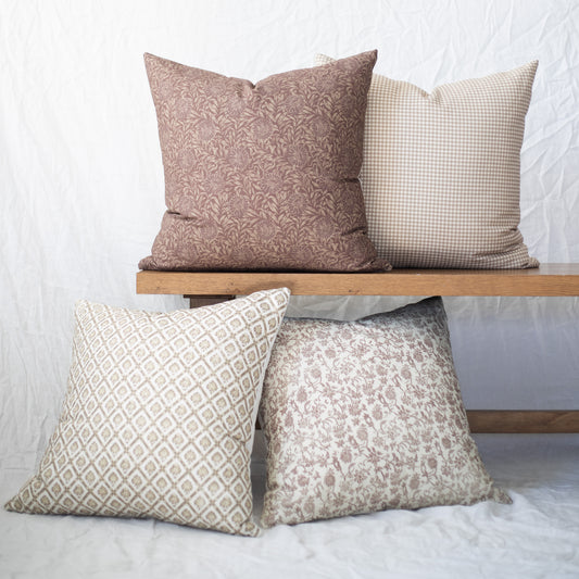Four decorative pillows in taupe and cream with floral, gingham, and geometric patterns displayed on wooden bench.