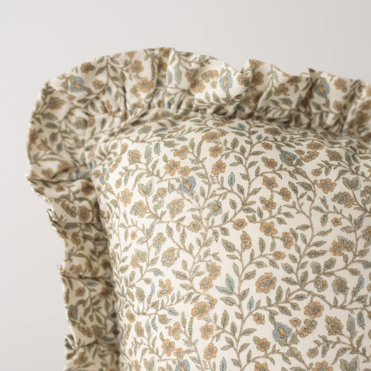 Cream pillow with ruffled edges and all-over floral pattern in tan, green, and blue tones.