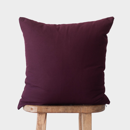 Deep merlot-colored pillow with square shape resting on a wooden stool against a light background.