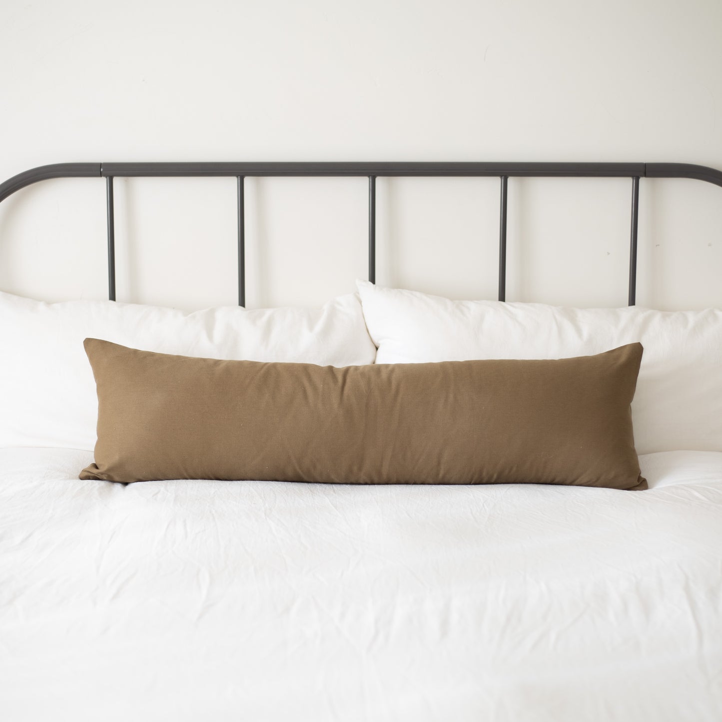 Tan lumbar pillow displayed on a bed with white pillows and metal headboard.
