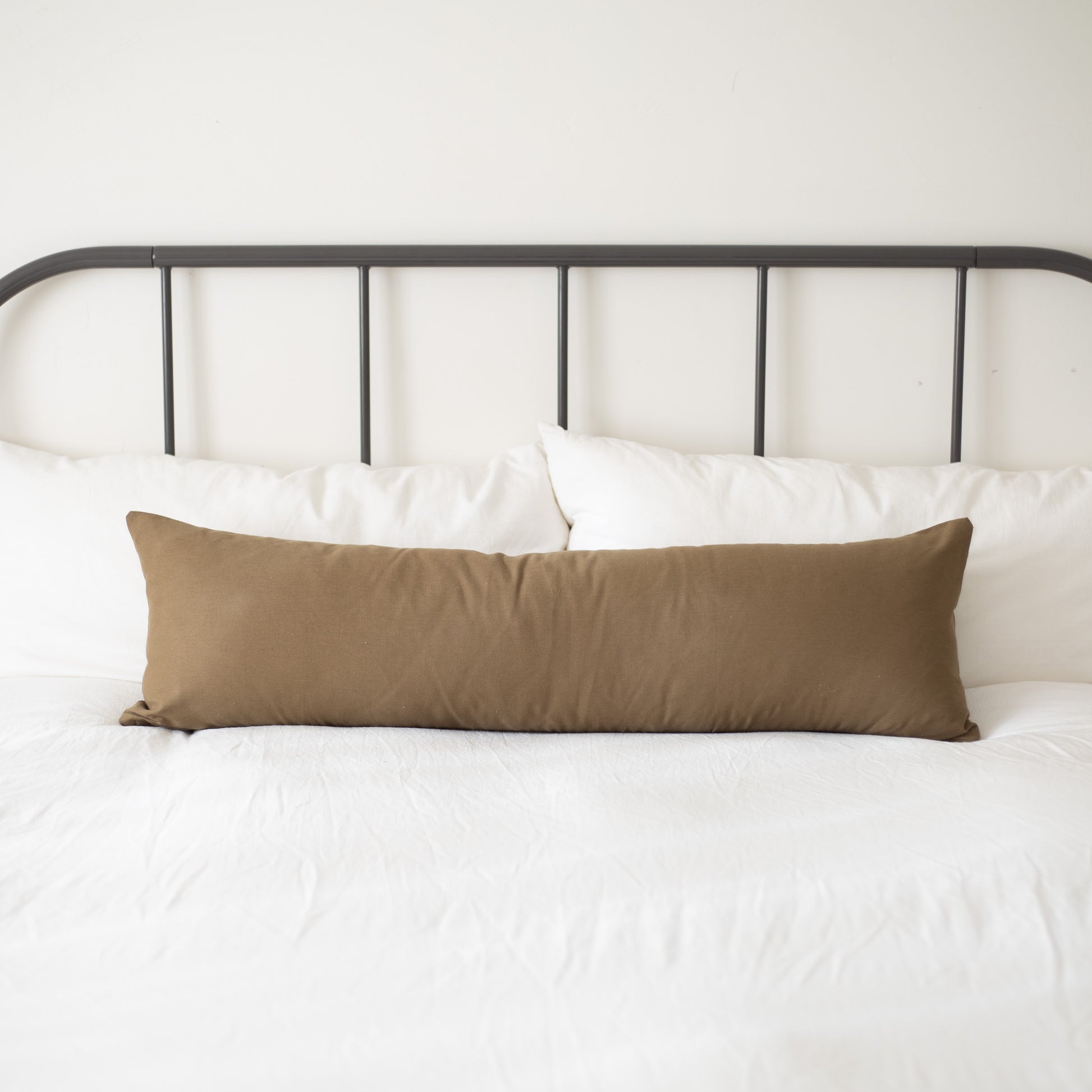 Tan lumbar pillow displayed on a bed with white pillows and metal headboard.