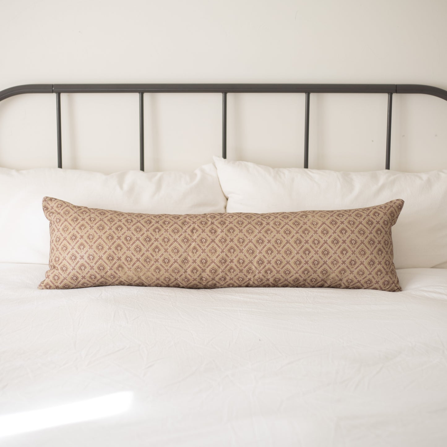 Tan lumbar pillow with geometric diamond pattern placed on white bedding against metal headboard.