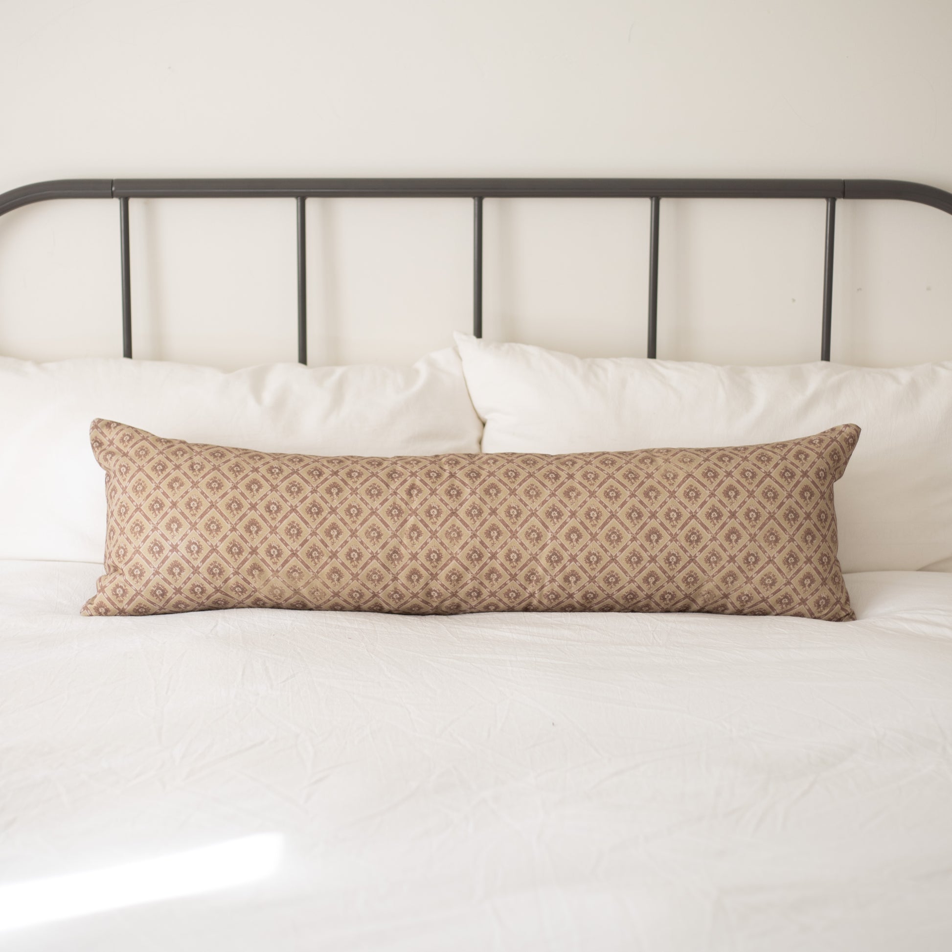 Tan lumbar pillow with geometric diamond pattern placed on white bedding against metal headboard.