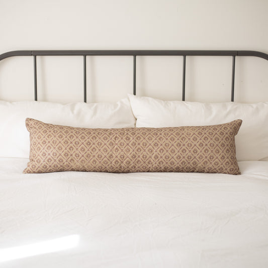 Tan lumbar pillow with geometric diamond pattern placed on white bedding against metal headboard.