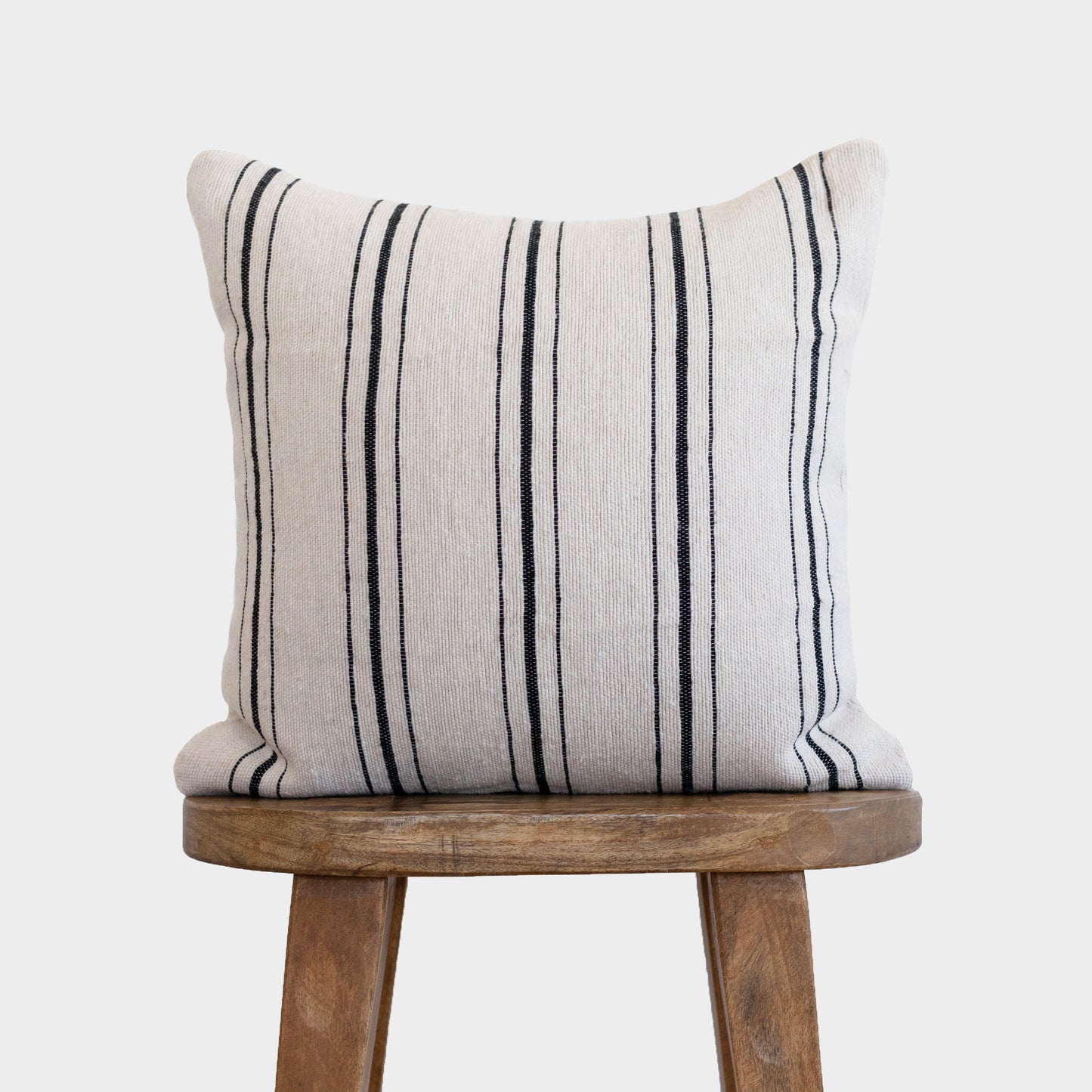 Cream linen pillow with black vertical stripes displayed on wooden stool.