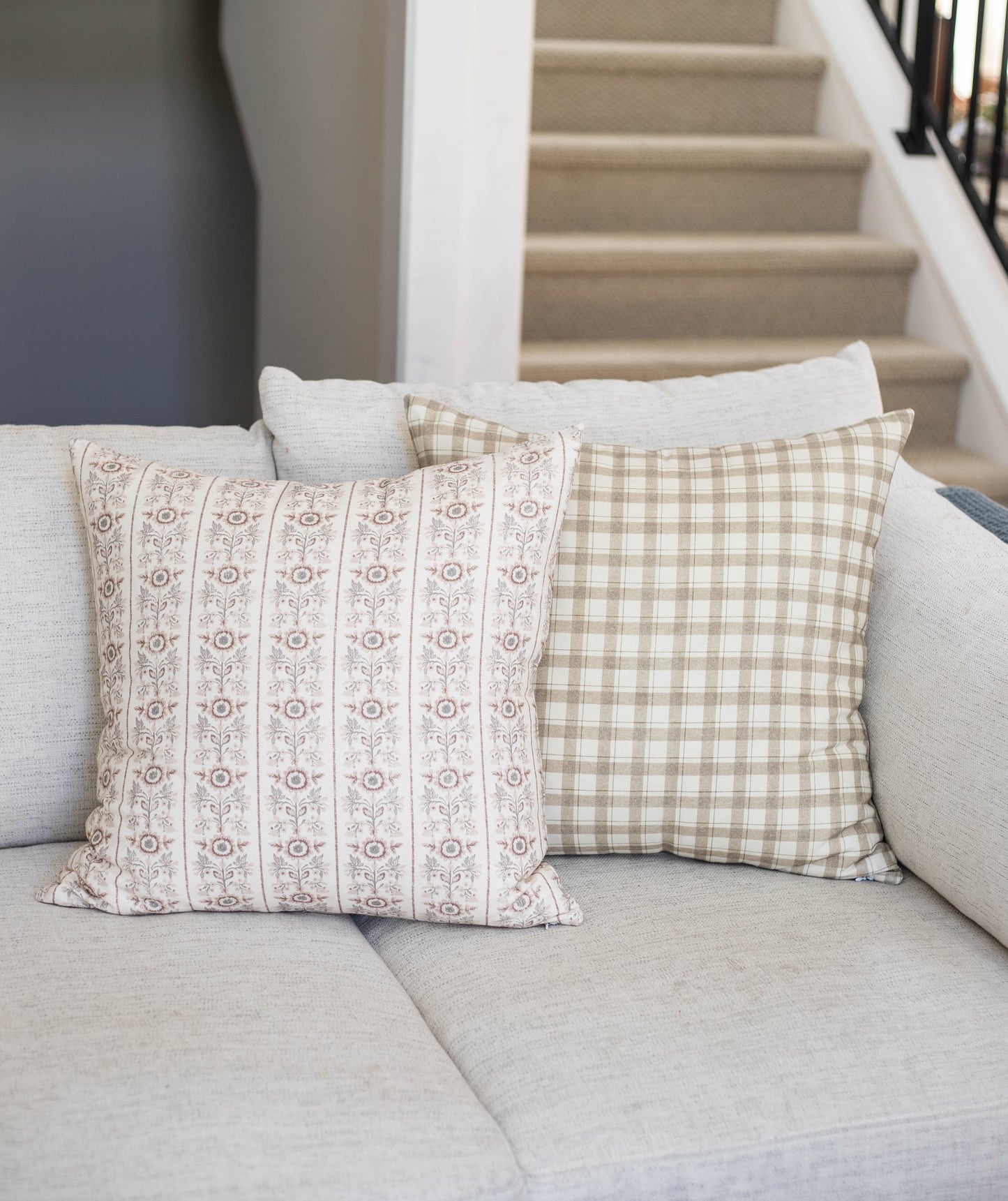 Four decorative pillows in cream, tan, and white with floral and gingham patterns arranged on a light gray sofa.