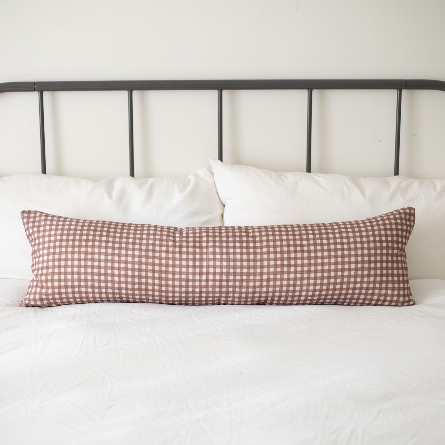 Rose and white gingham patterned lumbar pillow on a bed with white pillows and metal headboard.