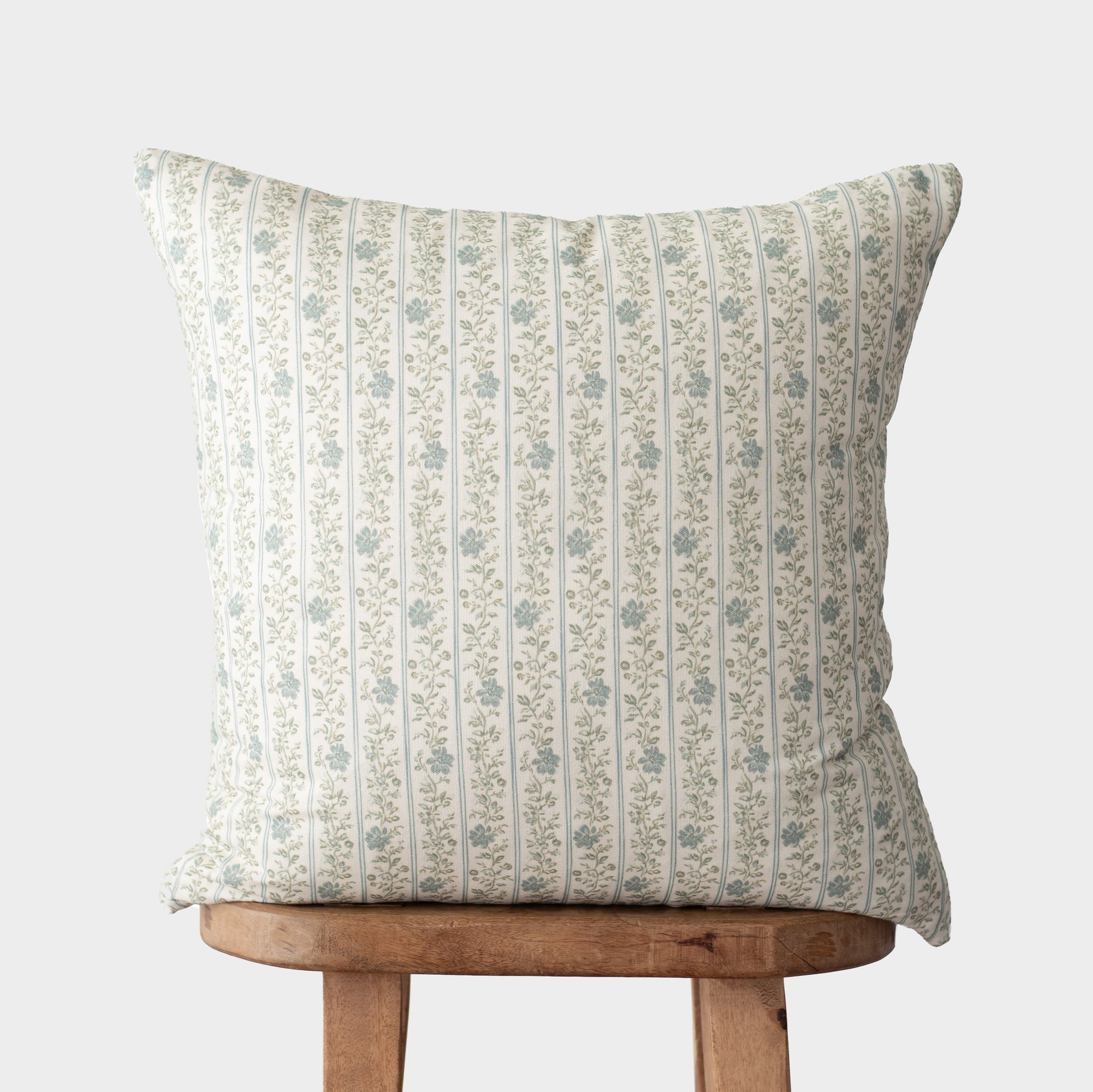 Cream pillow with vertical green stripes and small floral pattern, displayed on wooden stool.