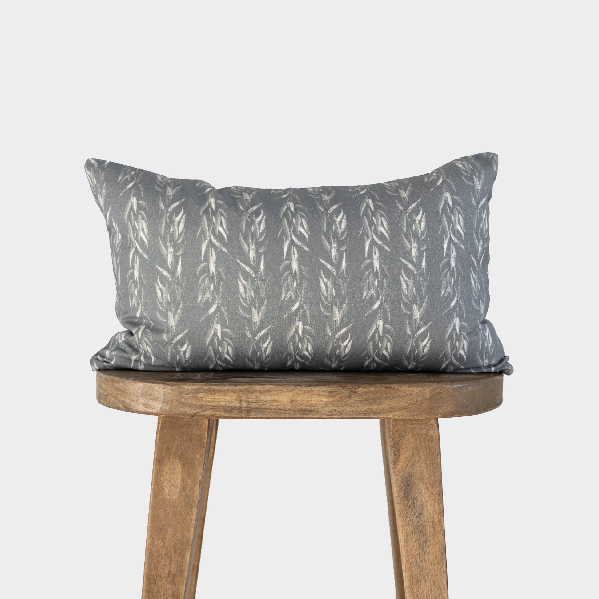 Rectangular lumbar pillow with sage green fabric featuring white leaf pattern, displayed on wooden stool.