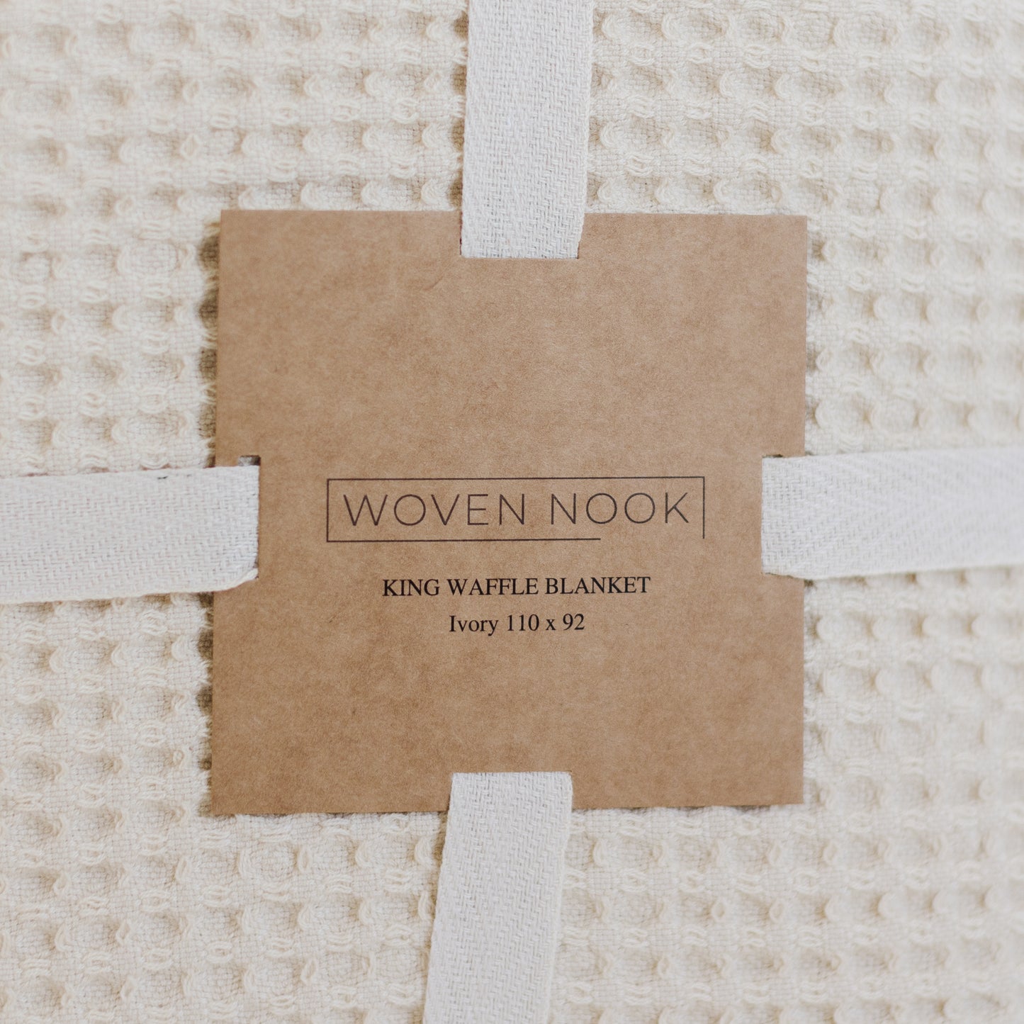 Ivory waffle textured blanket with white fabric straps and kraft paper product label.