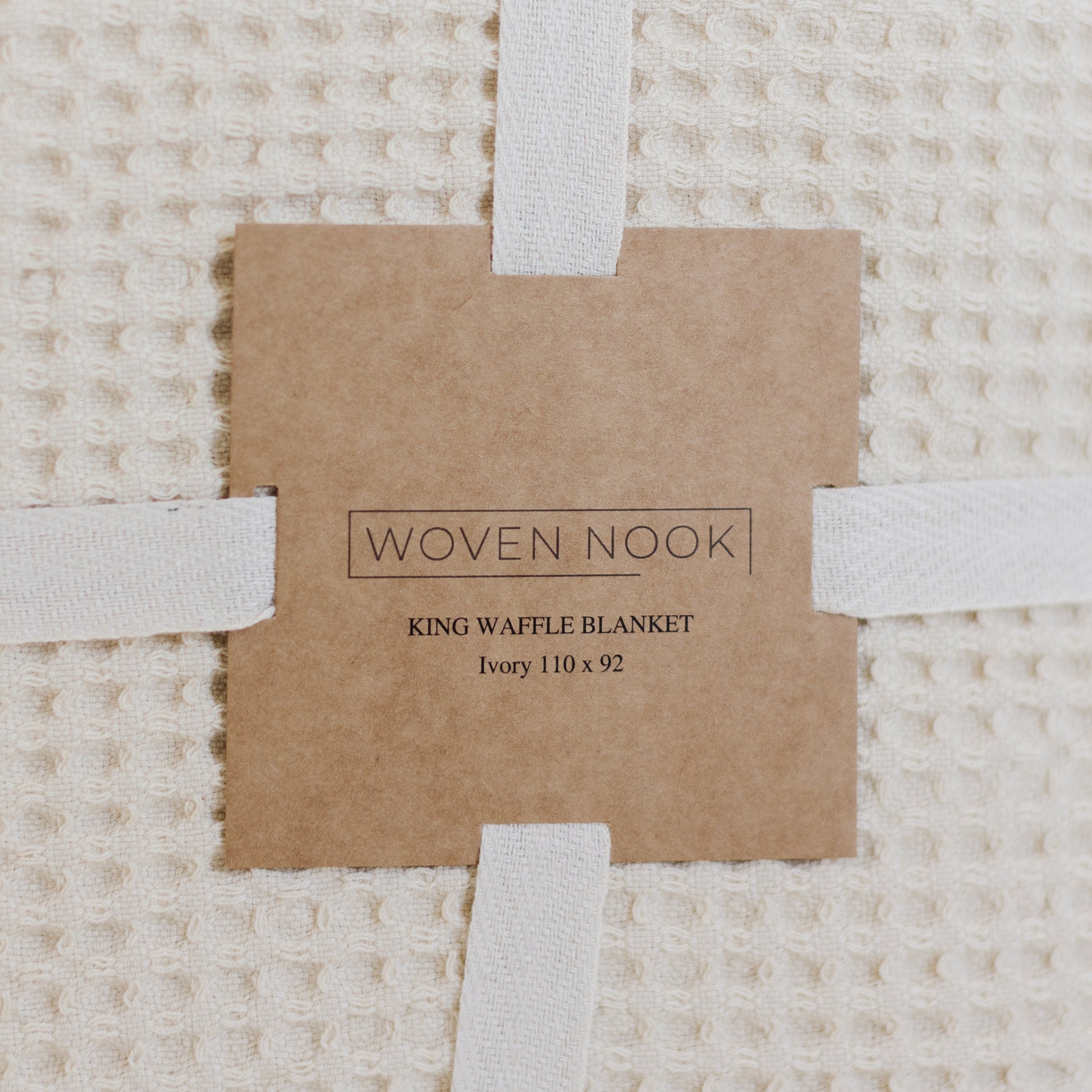Ivory waffle textured blanket with white fabric straps and kraft paper product label.