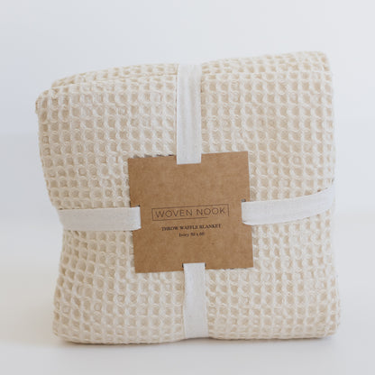 Ivory waffle blanket with textured grid pattern, rolled and wrapped with cream cotton band and kraft paper label.