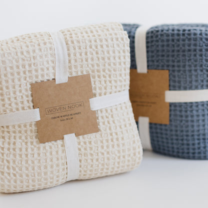 Rolled ivory waffle-textured blanket with kraft paper band and white fabric tie, displayed with a blue blanket.