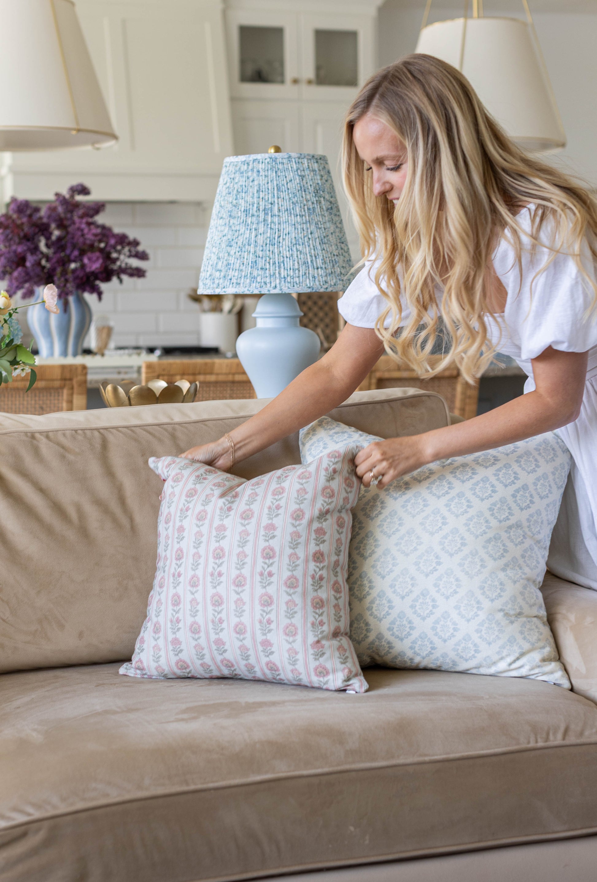 Woman arranging two decorative pillows with floral and geometric patterns on a beige sofa.