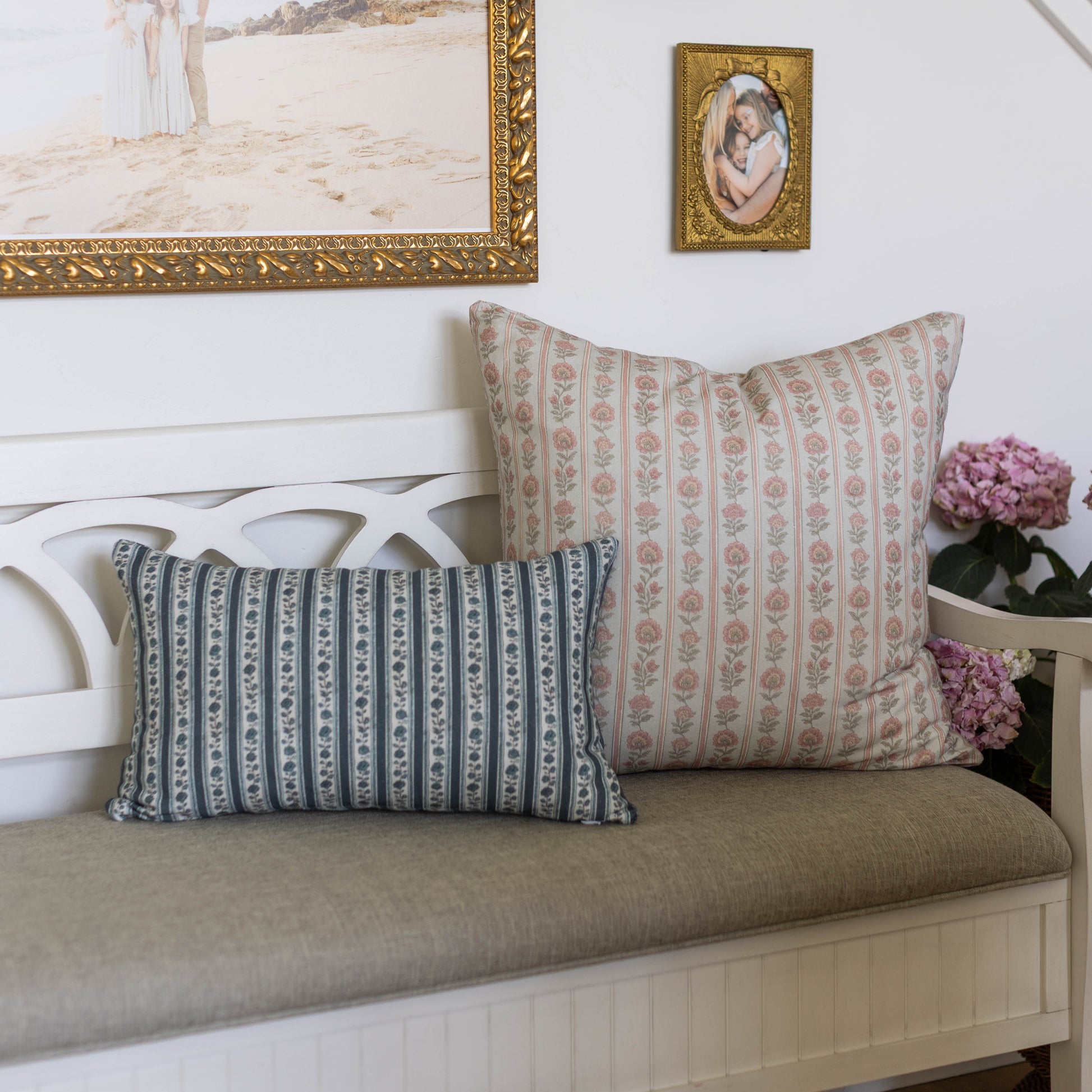 Decorative pillow with pink floral pattern on natural linen, displayed on white wooden bench with blue striped pillow.