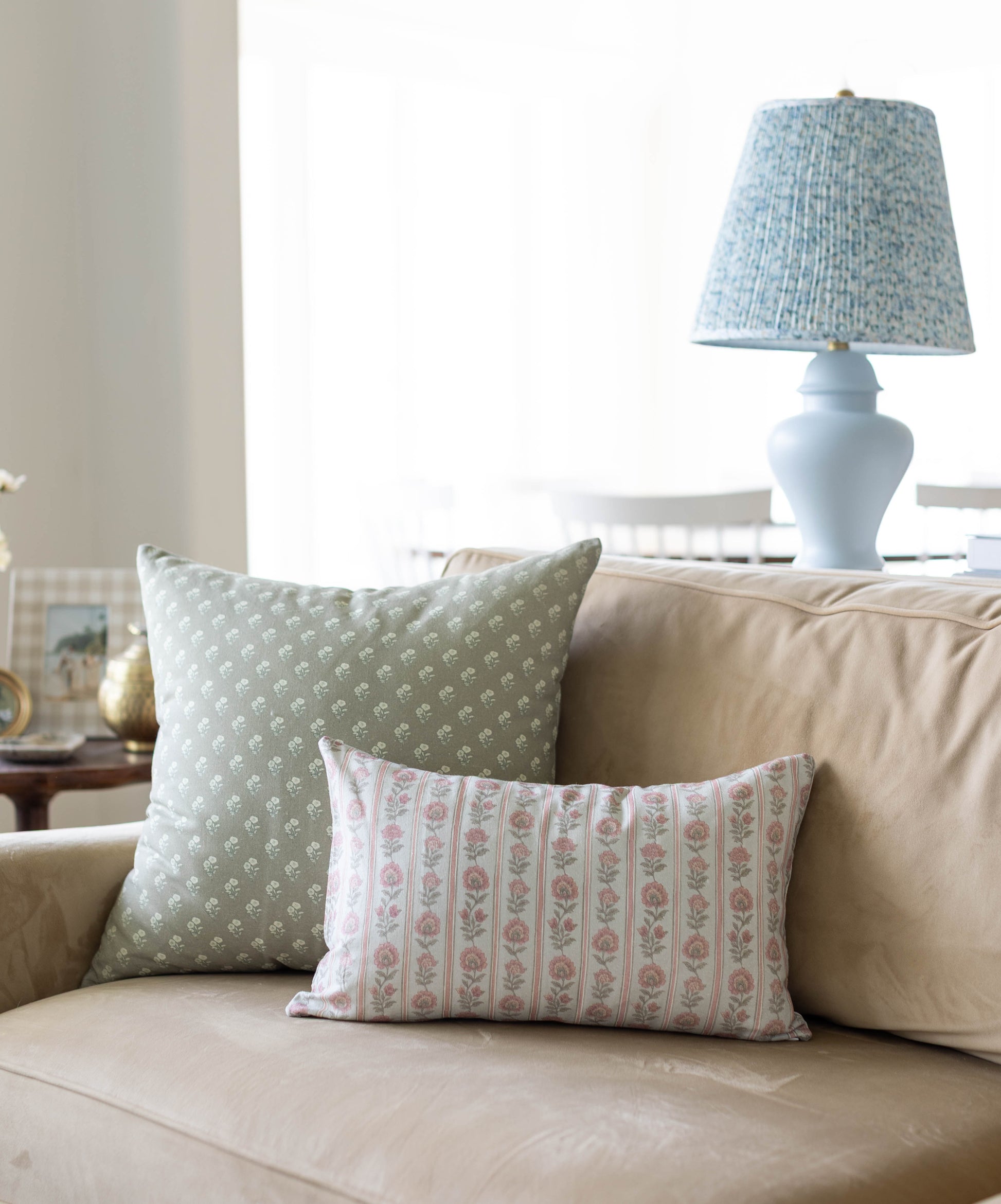 Light blue ceramic table lamp with pleated fabric shade on beige sofa with patterned pillows.