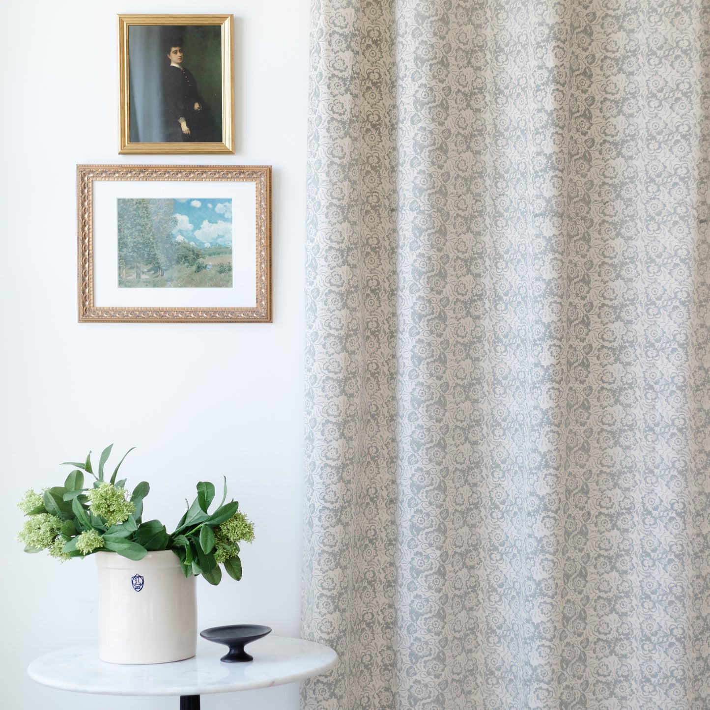 Two cream and taupe damask patterned curtain panels hanging at a window, displayed in a room with framed artwork and potted p