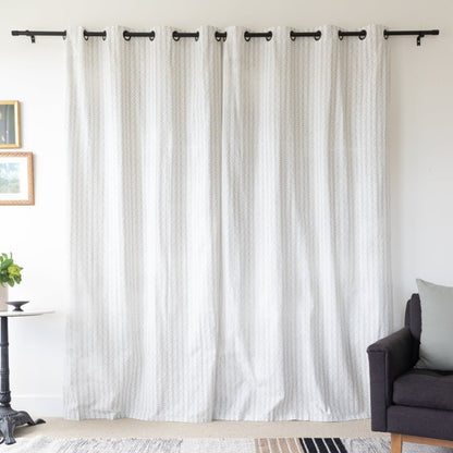 Two cream textured curtain panels with grommet tops hanging on a black rod in a living room setting.