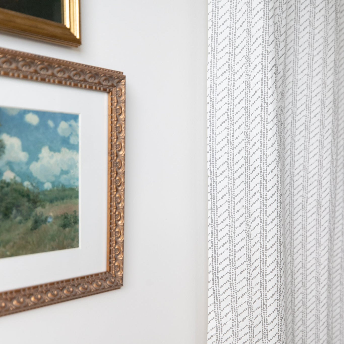 Two beige window curtains with diagonal textured pattern hanging next to framed landscape artwork.