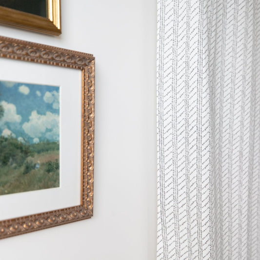 Two beige window curtains with diagonal textured pattern hanging next to framed landscape artwork.
