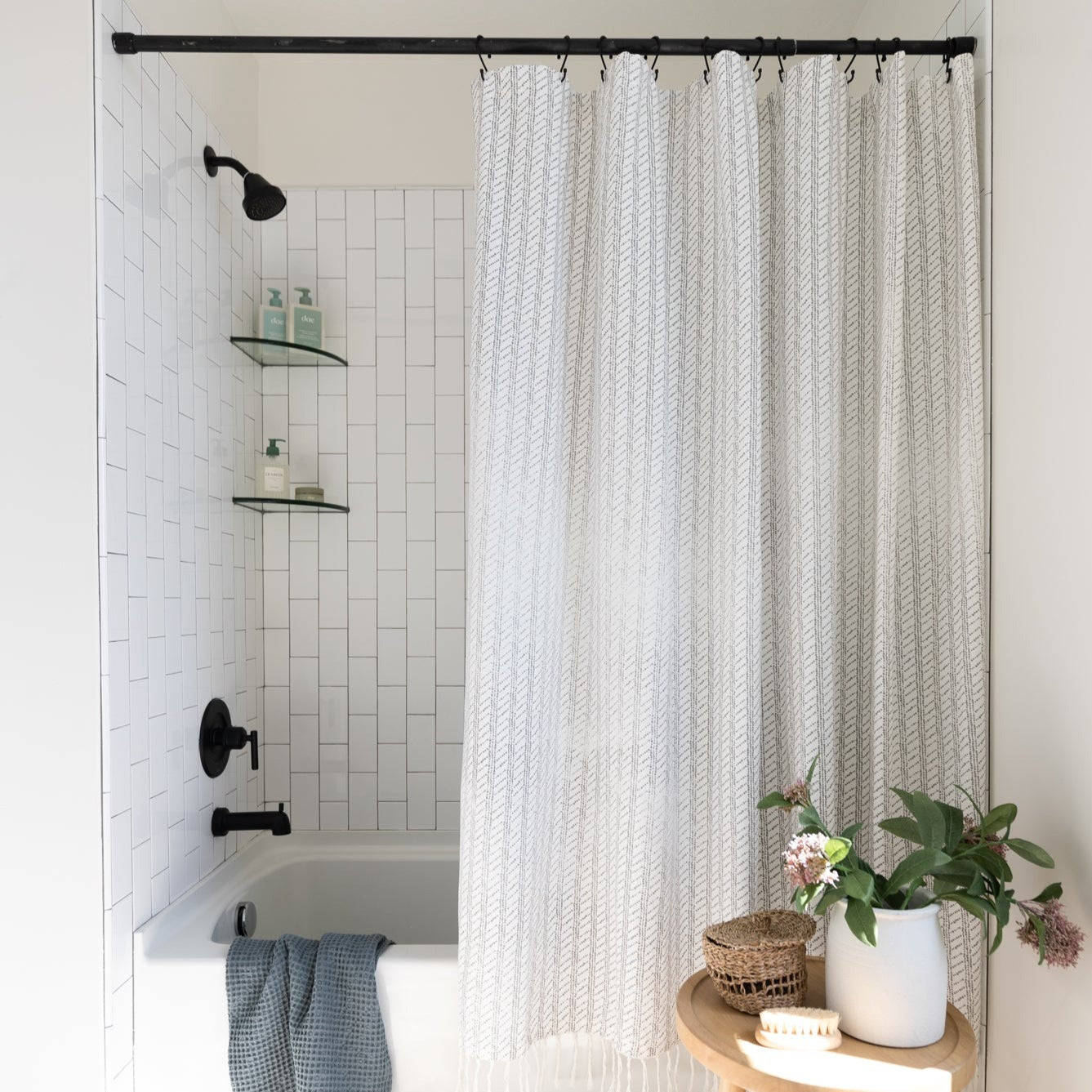 Cream textured shower curtain with vertical striped pattern hanging from black rod in white tiled bathroom.