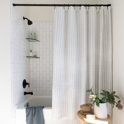 Cream textured shower curtain with vertical striped pattern hanging from black rod in white tiled bathroom.
