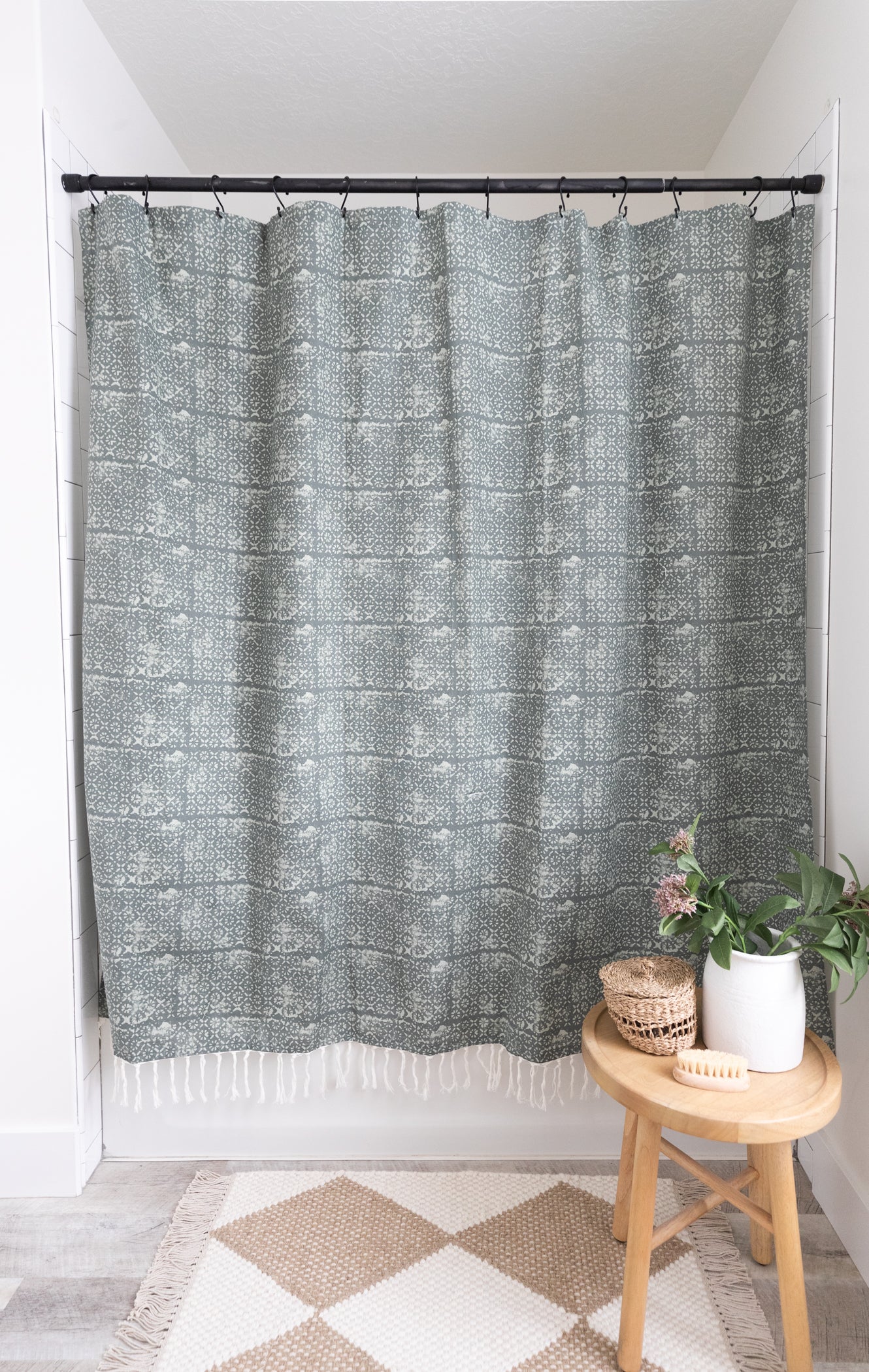 Gray patterned shower curtain with white fringe trim hung on black rod above white subway tile wall.