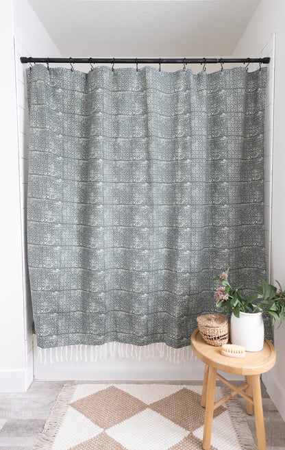 Gray patterned shower curtain with white fringe trim hung on black rod above white subway tile wall.