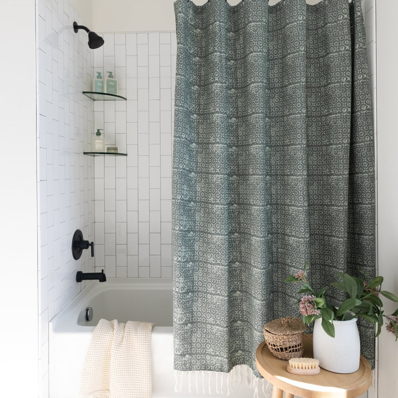 Green and white geometric patterned shower curtain hanging in a white tile bathroom with black fixtures.