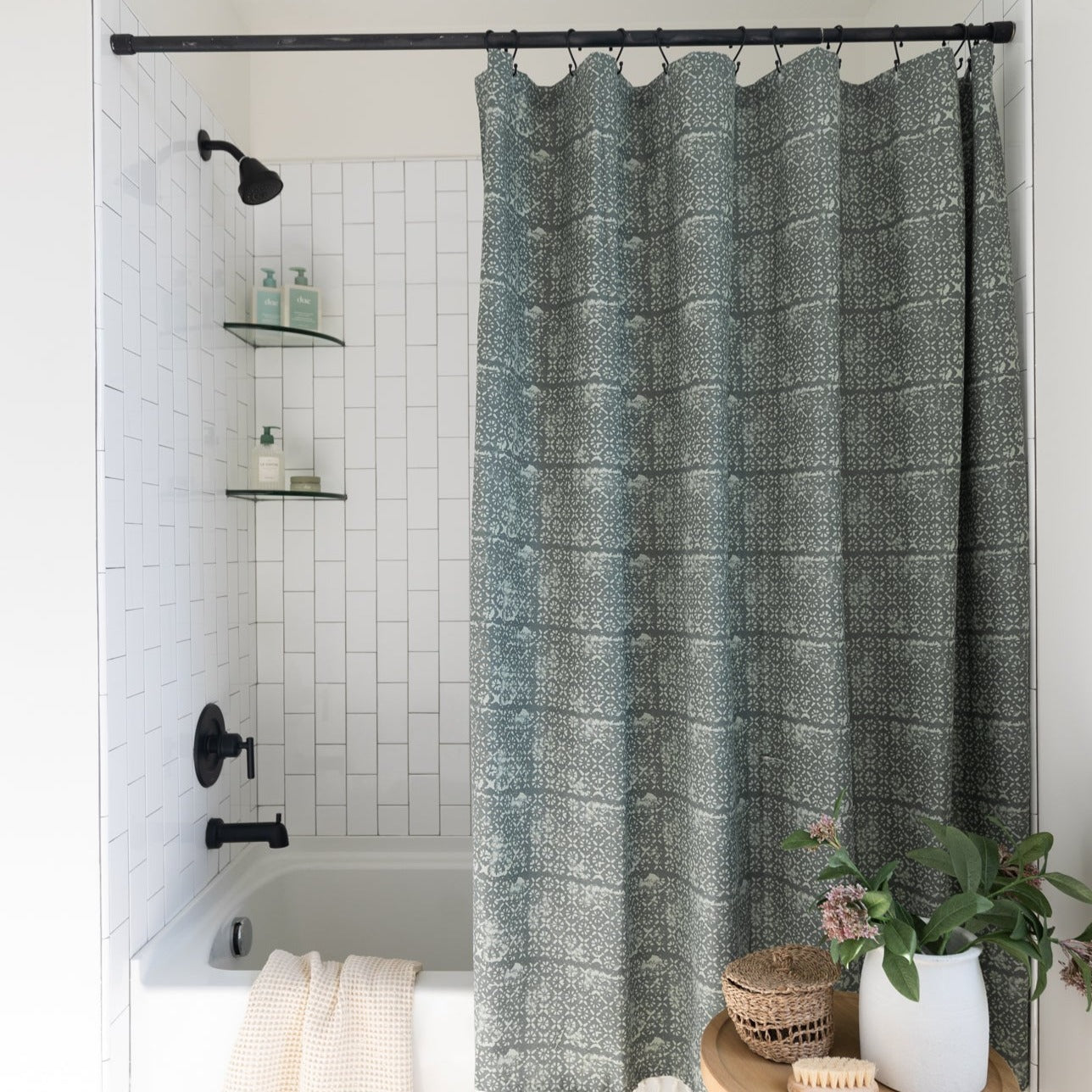 Sage green shower curtain with geometric damask pattern hung from black rod in white tile bathroom.